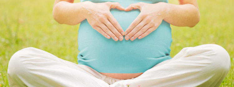 Foto einer schwangeren Frau, die im Gras sitzt und mit ihren auf den Bauch geklebten Händen ein Herz bildet