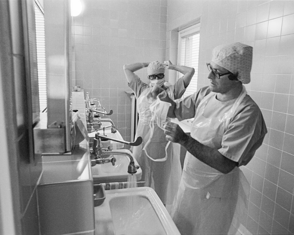 Bild aus de Wäschraim aus dem Operationszemmer, 2 Chirurgen wäschen hir Hänn fir sech virzebereeden ob eng Operatioun ( Dokter Guy Mandres, Dokter Norbert Weydert) . Si hunn eng speziell Kleedung un di nëmmen an deem Operatiounsbereich gedroen gëtt fir Infektiounen virzebeugen. Hir Hoer an de Mond a Nueseberaich sinn och ofgedeckt. Wärenddeem gëtt de Patient ob den Operatiounsdësch geluecht an d’Narkose gëtt ugefaangen. Den Oflaf vun de Gesten an de gudden Teamwork ass hei ganz wichteg fir d’Narkosezait zou kuerz wie méiglech ze halen. D’Foto ass aus dem Joer 1980 di Zäit gouf et an der Zitha 3 Operatiounssäll wou ënnert optimalen asepteschen Bedéngungen Operatiounen am Bauch, am Thorax, un de Schanken duerchfeiert gi sinn. Hei sinn och éischt kënstlech Gelenker agesat gi. Ugegliddert woren Lagerraim fir sterilt Material, e ganz groussen Sterelisator an Opbereedungsraim fir Material an Ekipementer, Openthaltzemmeren fir d’Mataarbechter. D’Bild ass vum Pol Aschmann Fotograph zu Lëtzebuerg.