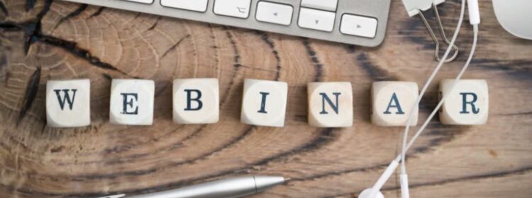 Foto von Würfeln, die mit den Buchstaben, die das Wort „Webinar“ bilden, vor einem Computer und Kopfhörern aufgereiht sind.